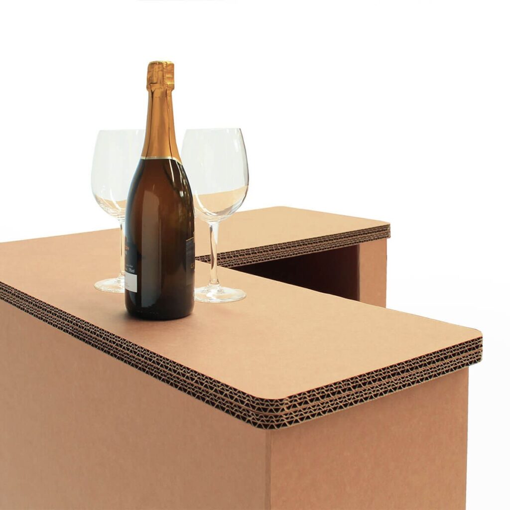 Comptoir en carton structuré avec plateau épais et compartiments intégrés, idéal pour organiser des espaces événementiels ou professionnels comme stands salons, bureaux ou salles de réception. Visible sur l'image : plateau cartonné renforcé et usage illustré par une bouteille et deux verres posés dessus - Le Meuble en Carton une marque du groupe Natur Design