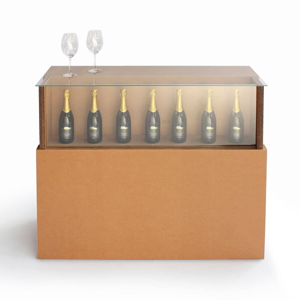 Comptoir en carton LOGAN E avec plateau transparent supportant des bouteilles de champagne alignées et deux verres à vin posés au-dessus, idéal pour l’aménagement de magasins, événements professionnels ou stands événementiels - Le Meuble en Carton une marque du groupe Natur Design