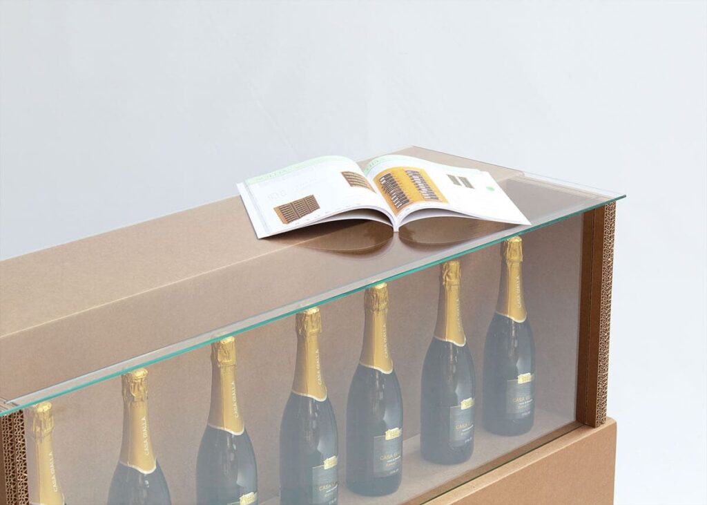 Comptoir en carton écologique avec plateau transparent pour présentation de bouteilles de champagne ou boissons premium, idéal pour stands événementiels et salons professionnels. Structure solide en panneaux cartonnés avec poignées métalliques et vitrage clair pour mise en valeur des produits - Le Meuble en Carton une marque du groupe Natur Design